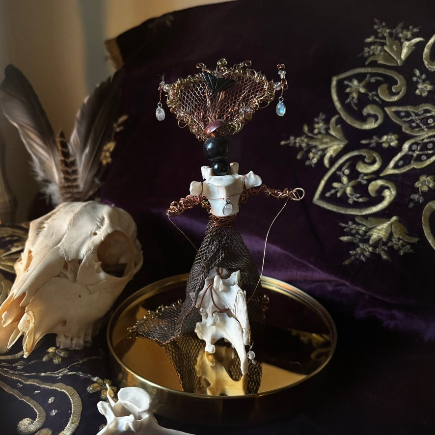 A gothic-style skeleton doll with a gold filigree hat and white bones, displayed on a gold tray next to a skull and dark purple embroidered fabric.